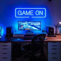 a desk with a computer and speakers on it in front of a neon sign that says game on