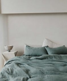 a bed with green sheets and pillows in a white room next to a vase on the wall