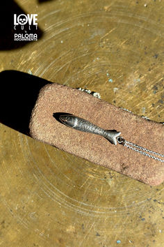 Silver fish pendant hanging from a silver chain head down laying on a brick-like stone on a brass surface. Sterling Silver Fish-shaped Necklace With Lobster Clasp, Antique Silver Hand Cast Sterling Silver Necklace, Hand Cast Antique Silver Sterling Silver Necklace, Hand Cast Sterling Silver Necklace In Antique Silver, Handmade Silver Fish-shaped Necklace, Sterling Silver Fish-shaped Jewelry Gift, Sterling Silver Fish-shaped Jewelry As Gift, Handmade Sterling Silver Fish-shaped Jewelry, Handmade Sterling Silver Fish Jewelry