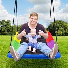 a man sitting on a rainbow colored swing with a baby in his lap and thumbs up