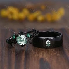 two black rings with green jewels on them sitting on a wooden table next to yellow flowers