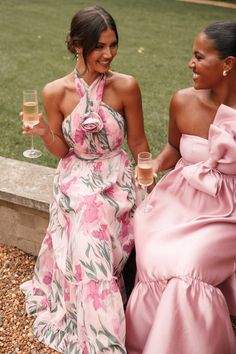 two women in pink dresses holding wine glasses and smiling at each other while sitting on the ground