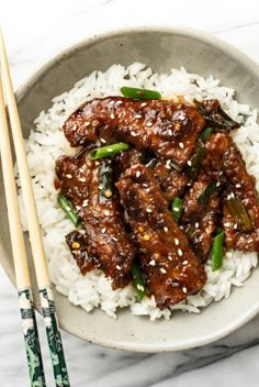 a bowl filled with rice and meat next to chopsticks on a marble table