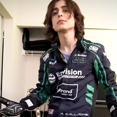 a man with long hair wearing a black and green racing suit standing in a room