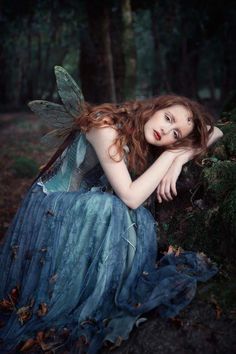 a woman with red hair and blue dress sitting on the ground in front of trees