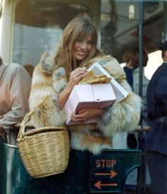 a woman in fur coat holding shopping bags and smiling while standing next to a bus