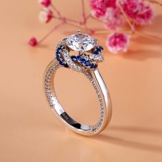 an engagement ring with blue and white stones on the side, sitting next to pink flowers