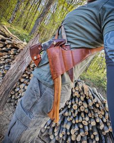 a man wearing a leather belt and carrying a knife in his back pocket, standing next to a pile of firewood