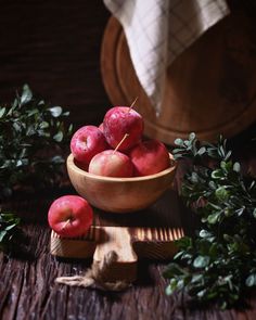 some apples are in a bowl on a wooden table next to greenery and a white towel