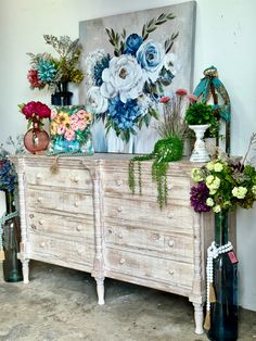 an old dresser with flowers and vases sitting on it's sideboard in front of a painting