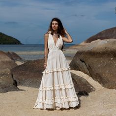 a woman in a white dress standing on the beach with rocks and water behind her