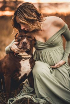 a woman in a long dress hugging her dog's face while sitting on the ground