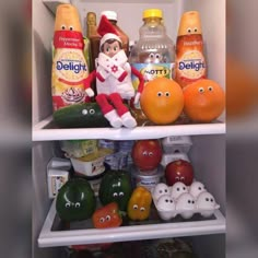 an elf is sitting on top of the refrigerator shelf filled with fruits and vegetables, including oranges