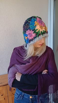 a woman with her arms crossed wearing a purple scarf and flowered knitted hat