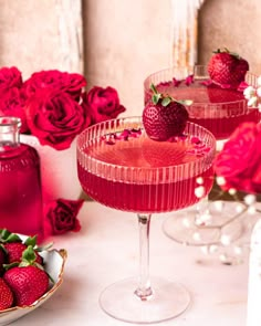 two wine glasses filled with pink liquid and strawberries on the table next to red roses