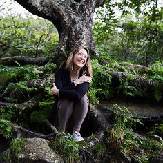 a woman sitting on the ground in front of a tree with roots growing out of it