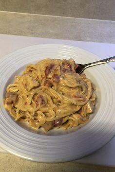 a white plate topped with pasta covered in sauce and bacon next to a silver spoon