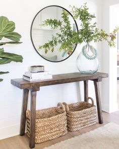 a wooden table topped with a vase filled with green plants next to a mirror and wicker baskets