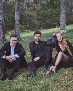 three people sitting on the ground in front of some trees, one wearing a suit and tie