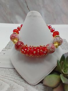 a red and gold beaded bracelet sitting on top of a white display stand next to succulents