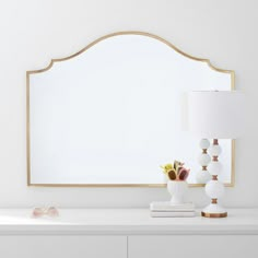 a white dresser topped with a mirror and vase filled with flowers next to a lamp