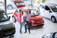 three women standing next to two cars in a showroom with the hood open and one woman raising her arms up