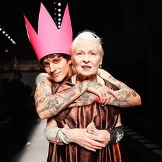 an older woman with tattoos and a crown on her head hugging another woman's shoulder