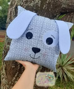 a hand holding up a pillow with a dog's face on it, in front of a tree