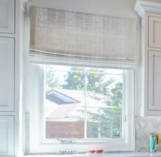 a kitchen with white cabinets and a window in the corner that has roman shades on it