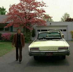 a man standing next to a white car in front of a tree with pink flowers