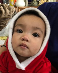 a baby wearing a red and white hoodie looking at the camera while sitting on a bed