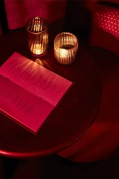 two lit candles are sitting on a table next to an open book and some glasses