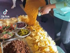 a woman is pouring sauce over food on a table with chips and olives in bowls