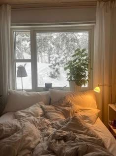 an unmade bed sitting in front of a window next to a night stand with a lamp on it