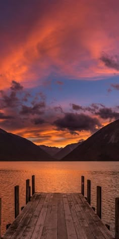 a wooden dock sitting on top of a lake under a colorful sky with clouds in the background