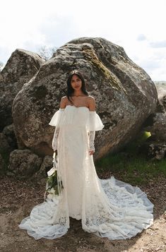 a woman standing in front of a large rock wearing a wedding dress with long sleeves