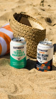 two cans of tiwa water sit in the sand next to a basket and towel