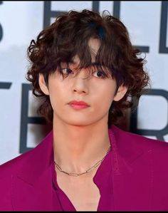 a man with curly hair wearing a purple suit and necklace at an awards event in front of a white background