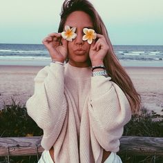 a woman holding two flowers in front of her face while sitting on a bench at the beach