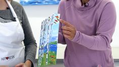 two women standing next to each other in front of paintings on the table and one is pointing at something