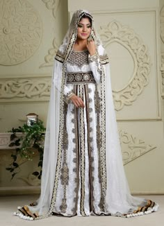 a woman wearing a white and gold wedding dress with a veil on her head, standing in