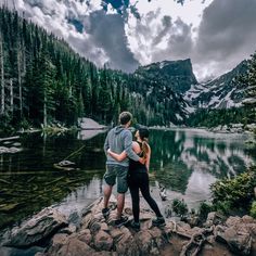 a man and woman standing next to each other on rocks near a lake with mountains in the background