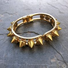 This stunning cuff is made of individually handset gold spikes in pronged settings, arranged around a hinge closure cuff. The oval shape fits comfortably on most wrists and is perfect for stacking or wearing solo. With its edgy design, this cuff will add a touch of rebellious glamour to any outfit. Whether you're dressing up for a night out or adding a little edge to your everyday look, the Helios Cuff is the perfect accessory for any occasion. DETAILS Bracelet Diameter: 7” Punk Metal Bangle Cuff Bracelet, Punk Style Metal Cuff Bangle Bracelet, Edgy Adjustable Gold Jewelry, Metal Studs Bracelets For Party, Party Bracelets With Metal Studs, Metal Bracelets With Studs For Party, Metal Party Bracelets With Studs, Trendy Spiked Jewelry For Parties, Trendy Metal Bracelets With Studs