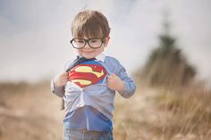 a young boy wearing glasses and holding a superman shirt on his chest, standing in the grass