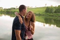 a man and woman standing next to each other holding beer bottles in front of a body of water