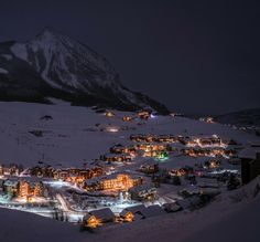 a town is lit up at night in the snow with mountains in the back ground