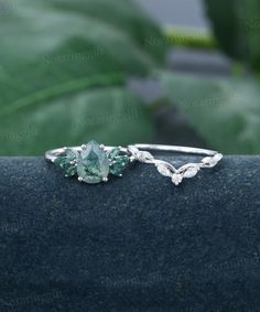 two rings with green stones on them sitting on top of a blue stone surface next to a plant