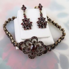 "Vibrant red Bohemian garnets sparkle on this vintage bracelet and earring set. The settings and are composed of yellow gold vermeil over 900 silver (a slightly lower purity of silver than sterling or 925). The faceted gemstones are a deep saturated red color with flashes of brighter red. They are of varying shape, cuts and 0.9mm to 7.6mm sizes. The bracelet has a flower shaped focal piece with a large oval 7.6mm x 5.9mm garnet at its center. A halo of six bezel-set 1.5mm stones surround it. The Elegant Handmade Brass Jewelry Sets, Elegant Hand-set Brass Jewelry, Elegant Hand Set Brass Jewelry, Evening Ruby Jewelry With Matching Earrings, Evening Ruby Jewelry Set With Matching Earrings, Evening Ruby Jewelry, Round Shape, Victorian Oval Jewelry For Evening, Victorian Jewelry With Oval Matching Earrings, Victorian Jewelry Bracelet With Jewels