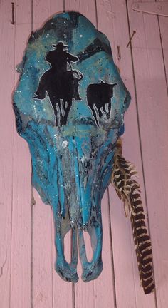 a cow skull with two cowboys on it and a feather in the foreground, against a pink painted wood background