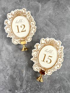 two gold and white wedding table numbers on top of a gray stone surface with golden bells